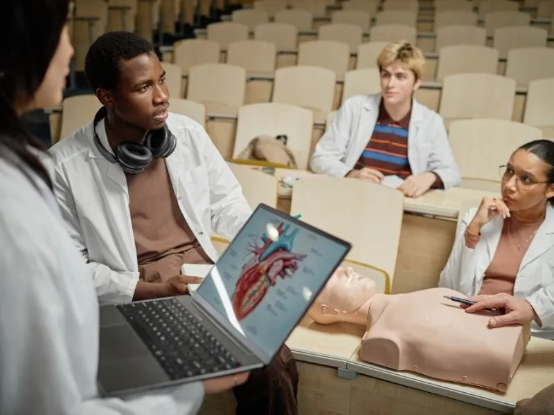 Grupo de estudantes de medicina em aula prática na Argentina, utilizando simulador realístico e laptop com anatomia cardíaca.